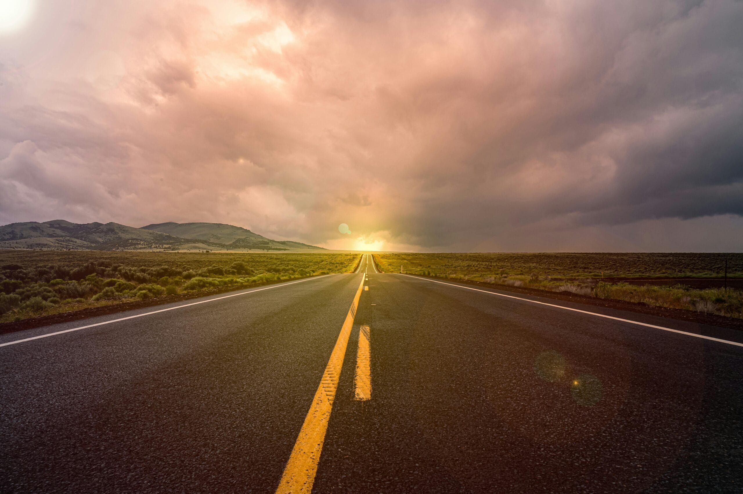 Open road stretching toward the horizon under a dramatic cloudy sky, with distant hills and evening sunlight in the background.